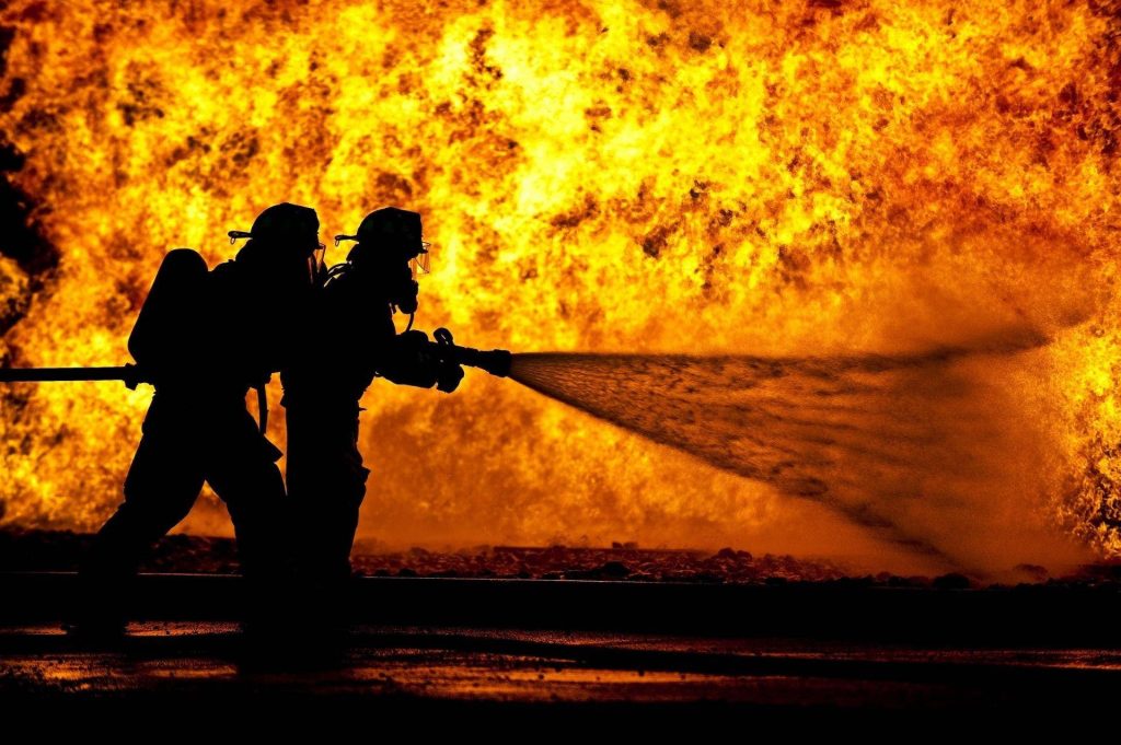 Two firefighters battle blazing fire spraying water heroic action intense wildfire wallpaper background