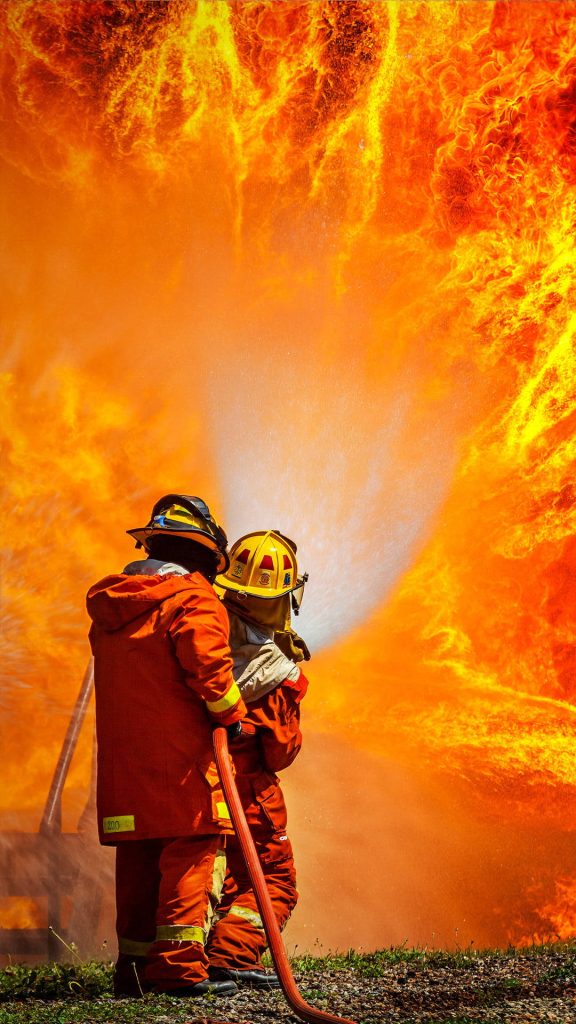 Two firefighters in protective gear battling a massive blazing inferno with water spray Wallpaper