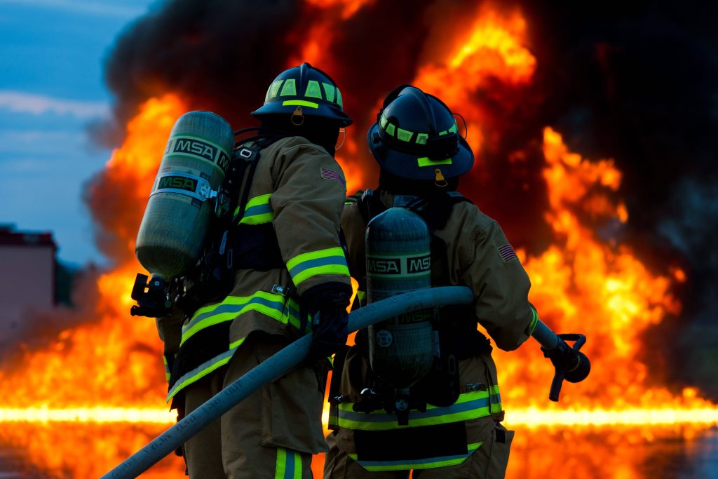 Two firefighters in full gear with oxygen tanks face a huge blazing orange wildfire background Wallpaper