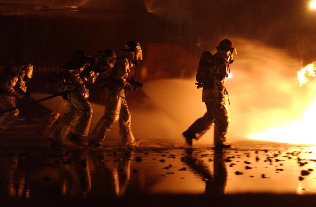 Firefighters in reflective gear march toward large blazing fire in smoky intense cinematic scene Wallpaper