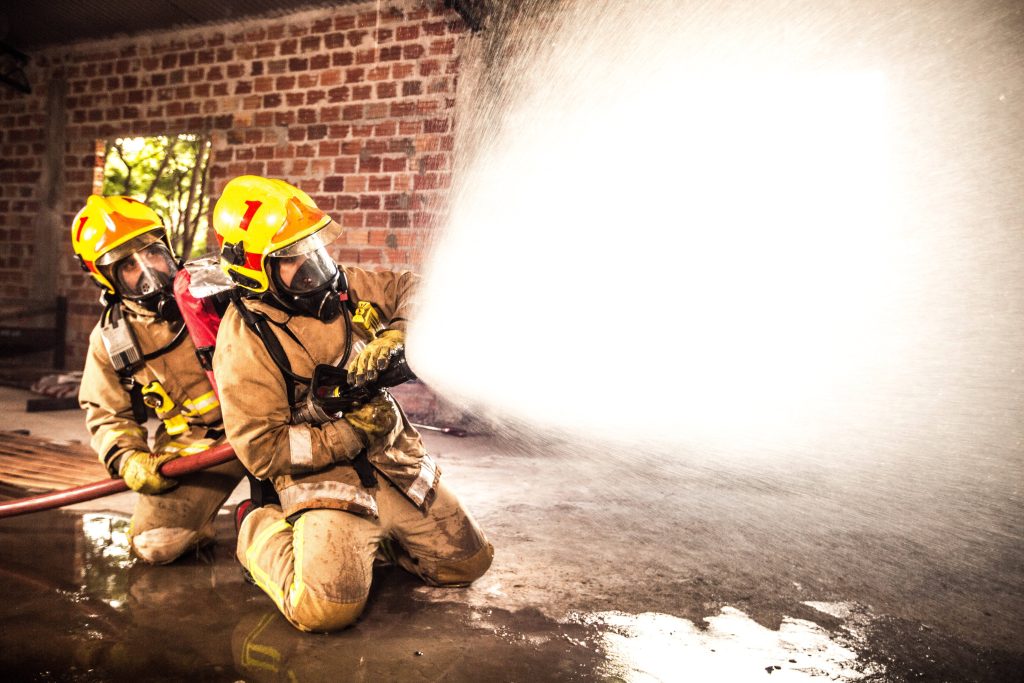 Two firefighters in full gear kneeling and using a high-pressure hose in industrial building Wallpaper