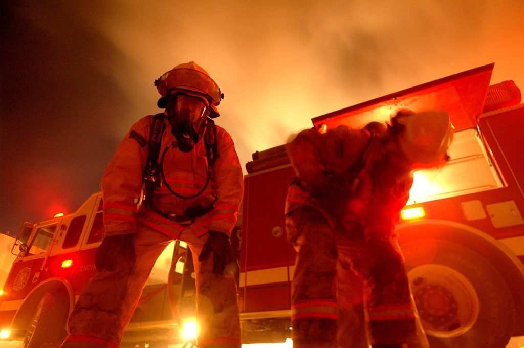 Two firefighters in full gear stand before fire engine in smoky orange glow emergency scene Wallpaper