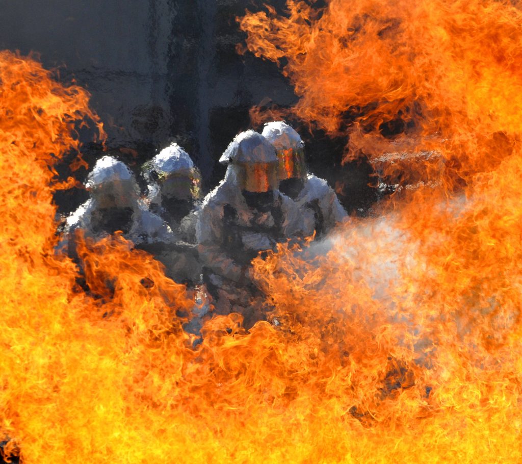 Four firefighters in aluminized suits battling intense orange and yellow flames wallpaper background