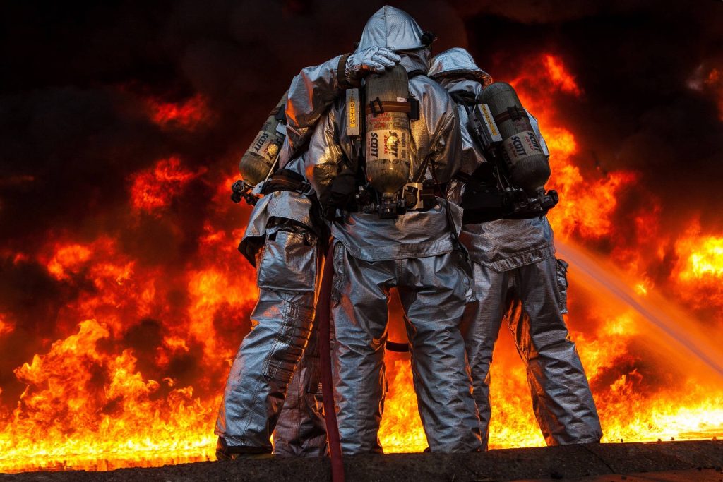 Three firefighters in silver reflective suits with Scott Air-Paks face a blazing orange fire background Wallpaper