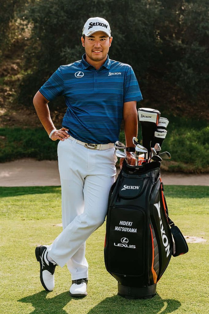 Professional golfer Hideki Matsuyama in blue polo leans on custom Srixon golf bag outdoors Wallpaper