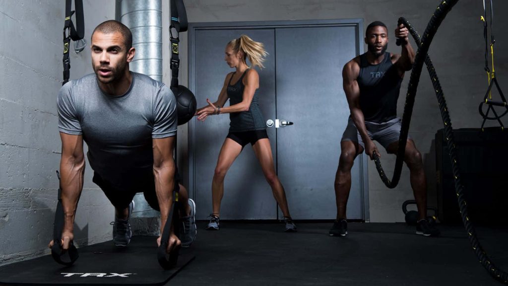 Three athletes training intensely with TRX straps and battle ropes in industrial gym background Wallpaper