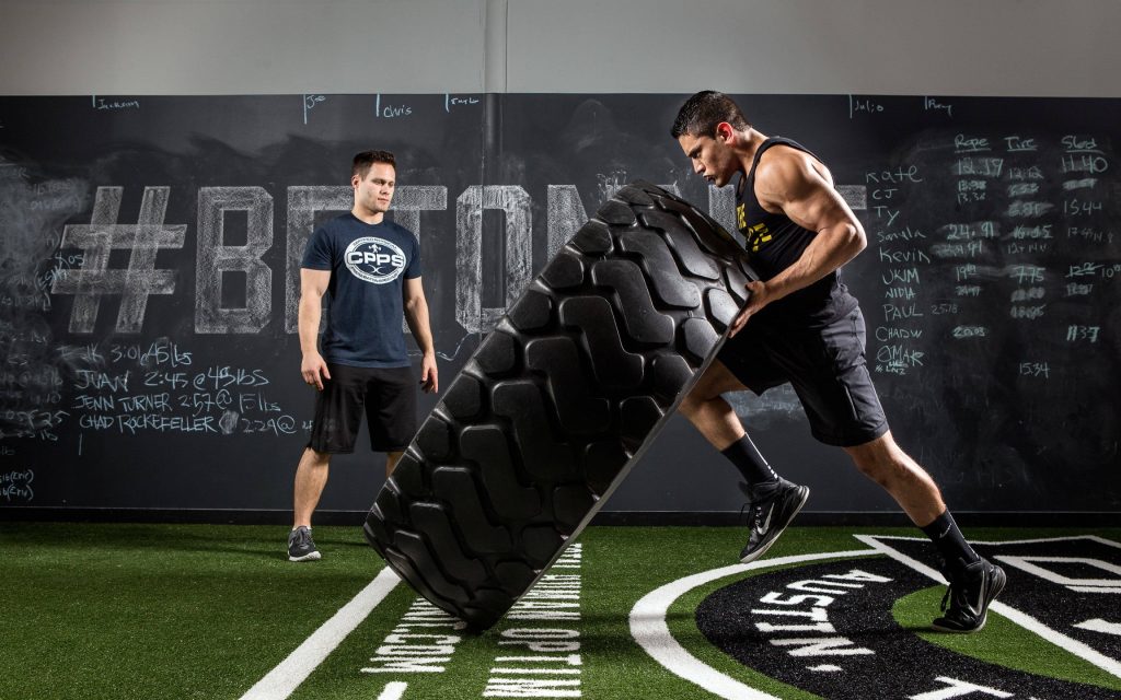 Focused athlete flipping heavy tire in gym with trainer and chalkboard workout leaderboard background Wallpaper