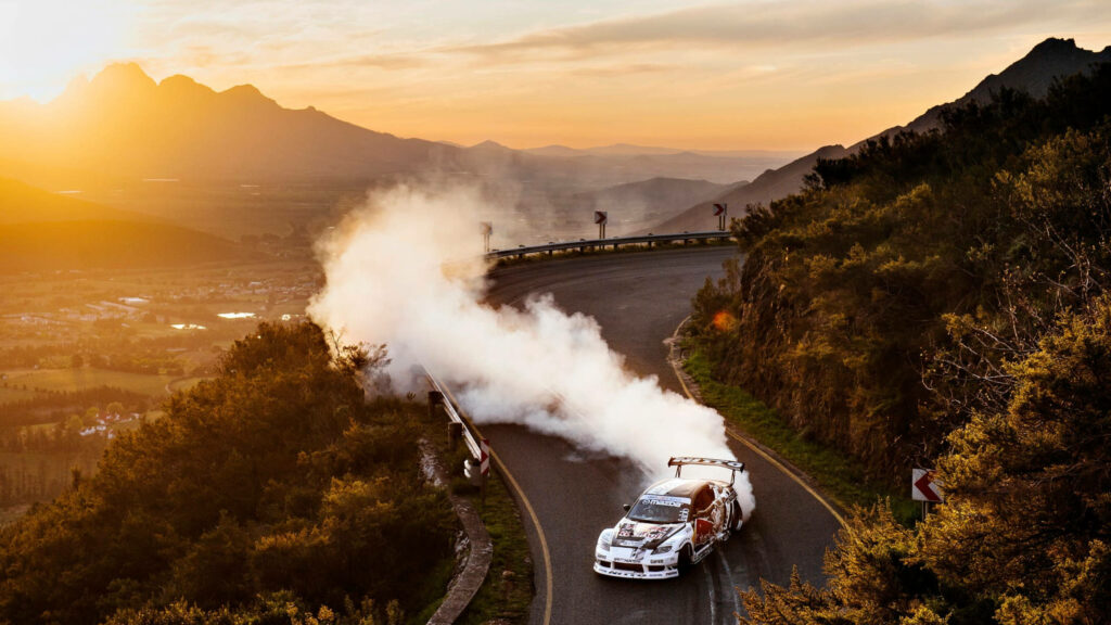 High-performance racing car drifting on sharp mountain road with smoke and sunset backdrop Wallpaper