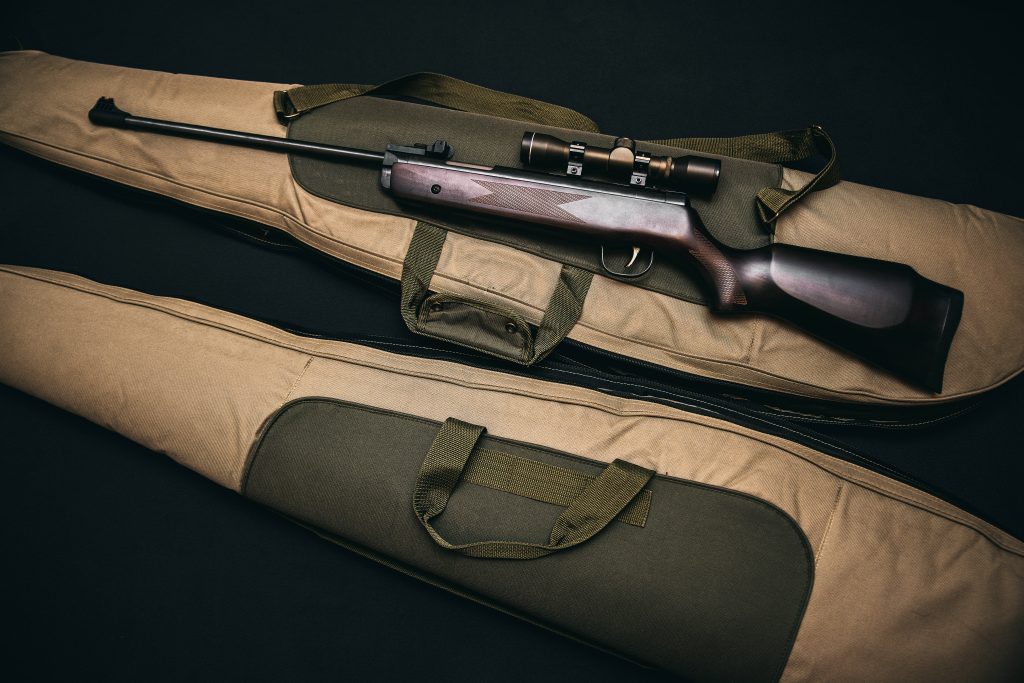 Scoped rifle with wooden stock on tan olive green padded case dark minimalist background Wallpaper