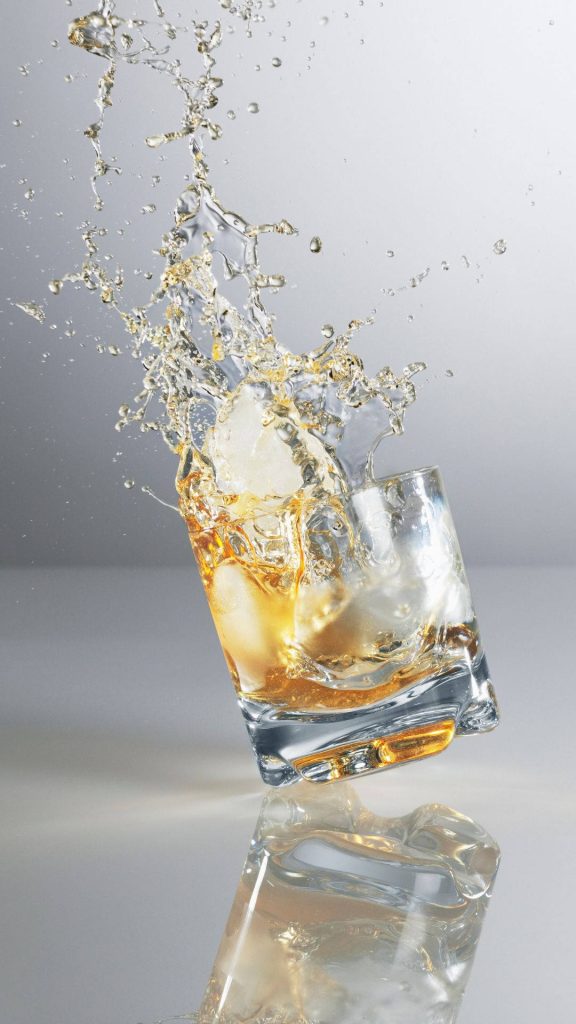 High-speed splash of amber whiskey in tilted rocks glass with ice on minimalist background wallpaper