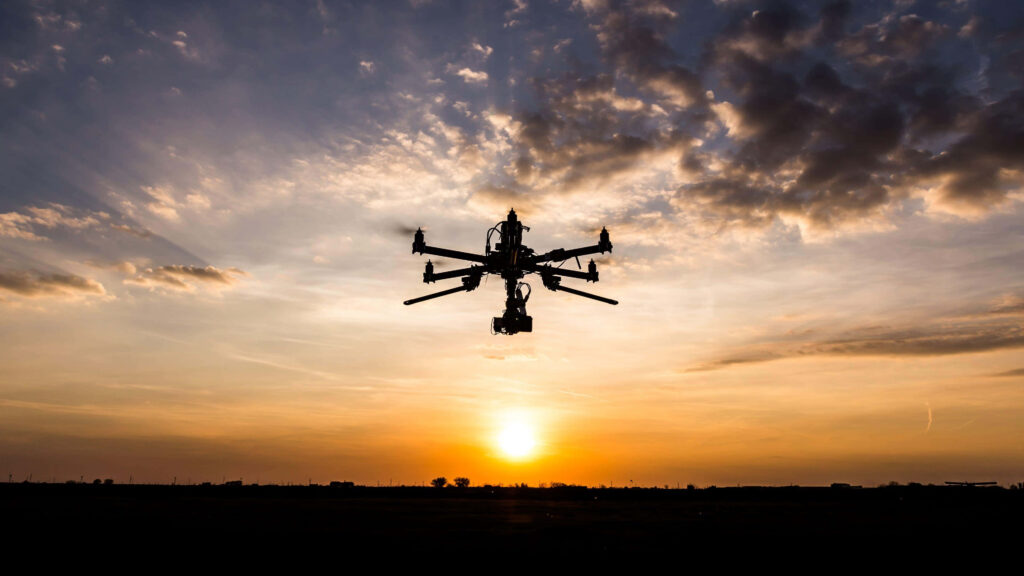 High tech drone with multiple rotors and camera silhouetted against vibrant sunset sky background Wallpaper