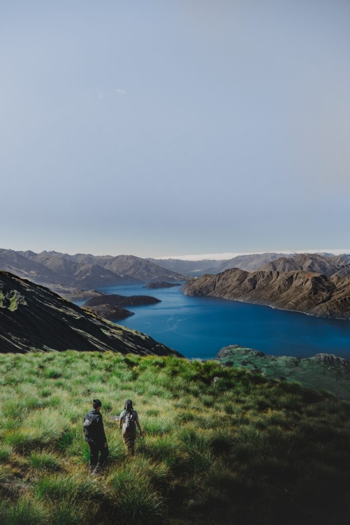 Two hikers on green hill overlook blue lake and rugged mountains under clear sky landscape wallpaper