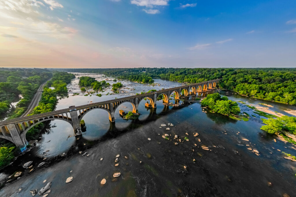 Historic arched bridge over calm river with lush green trees and clear blue sky wallpaper background