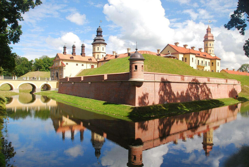 Historic Belarus red brick castle reflected in lake with blue sky white clouds and greenery wallpaper
