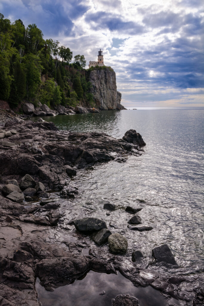Historic lighthouse on rugged cliff overlooking calm sea under dramatic clouds with green trees background Wallpaper