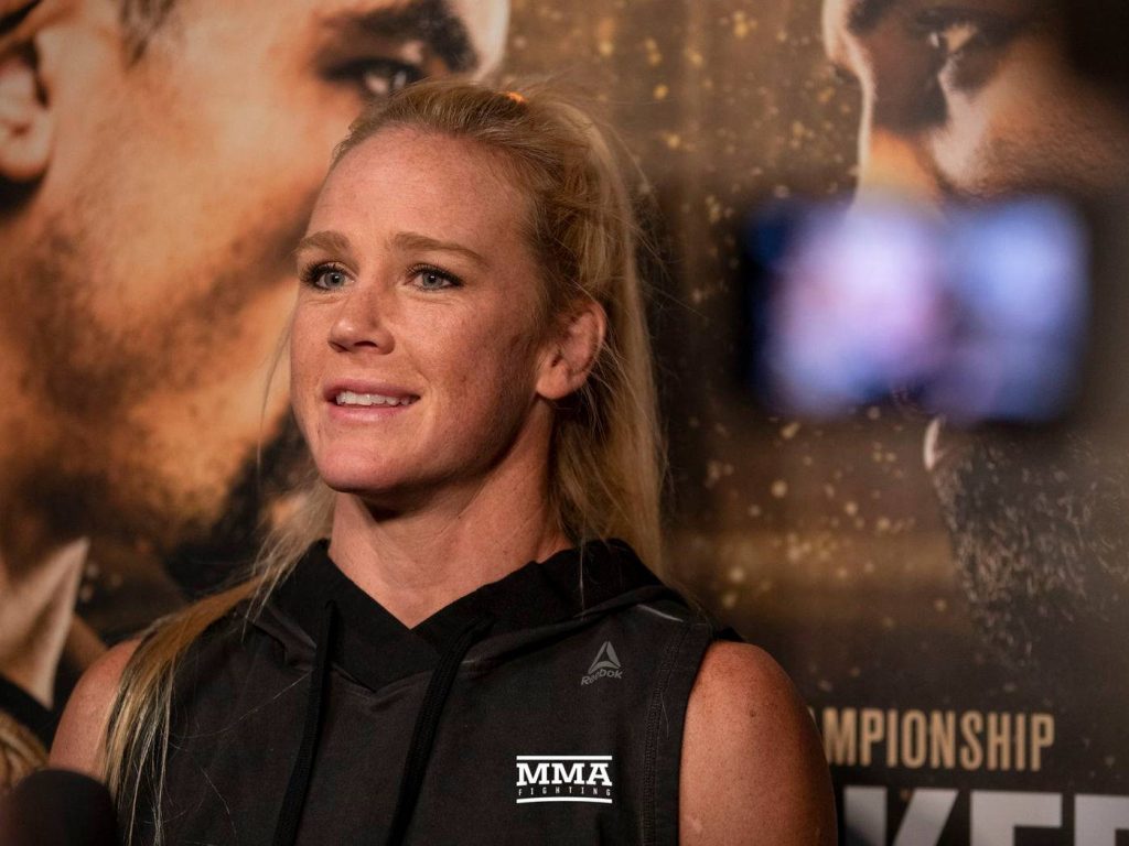 UFC fighter Holly Holm in black Reebok MMA top smiling before golden press backdrop wallpaper