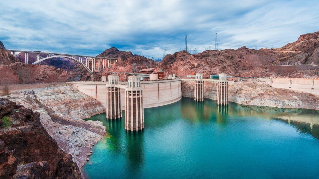 Hoover Dam with turquoise water rugged cliffs and Mike O'Callaghan Bridge under cloudy sky Wallpaper