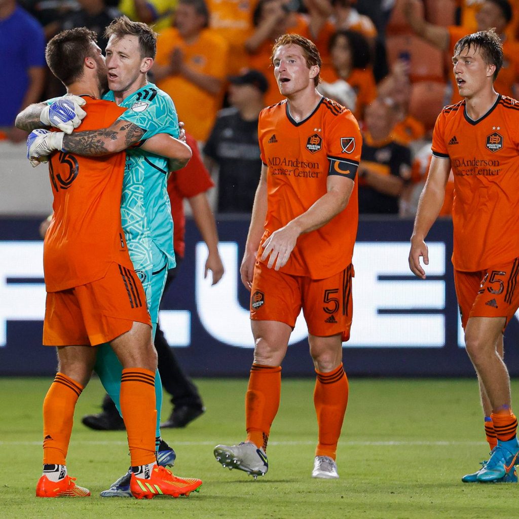 Houston Dynamo FC players in orange jerseys celebrate with goalkeeper in teal under stadium lights Wallpaper