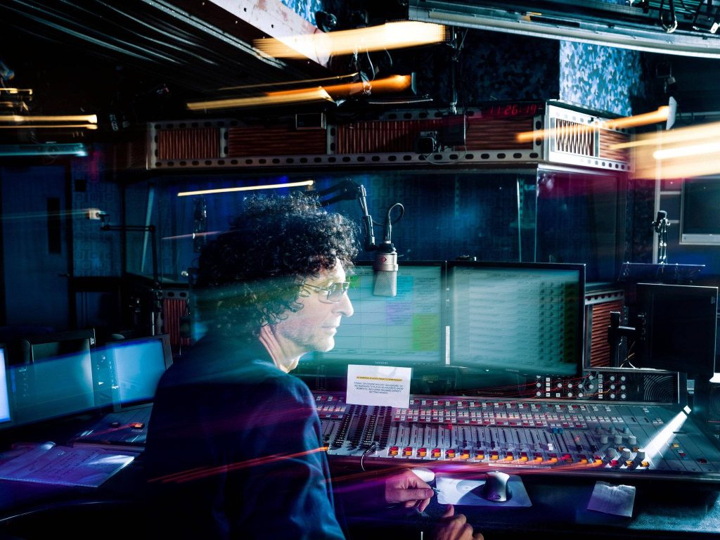 Howard Stern in radio studio with blue lighting mixing console and dynamic light trails wallpaper