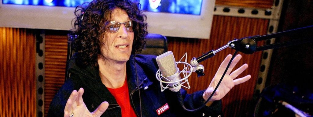 Howard Stern in broadcast studio with microphone curly hair tinted glasses acoustic panels wallpaper