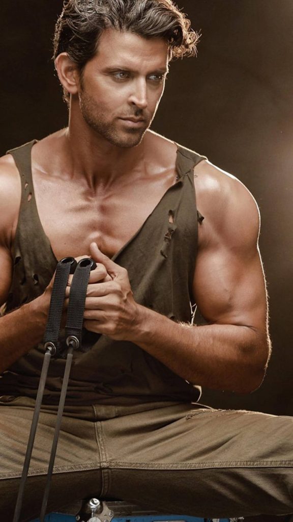 Hrithik Roshan muscular fitness wallpaper with warm lighting and rugged tank top holding resistance bands