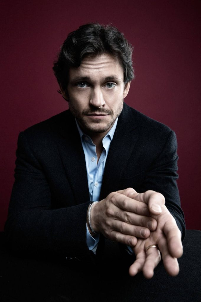 Hugh Dancy portrait intense gaze clasped hands dark suit light blue shirt maroon background Wallpaper