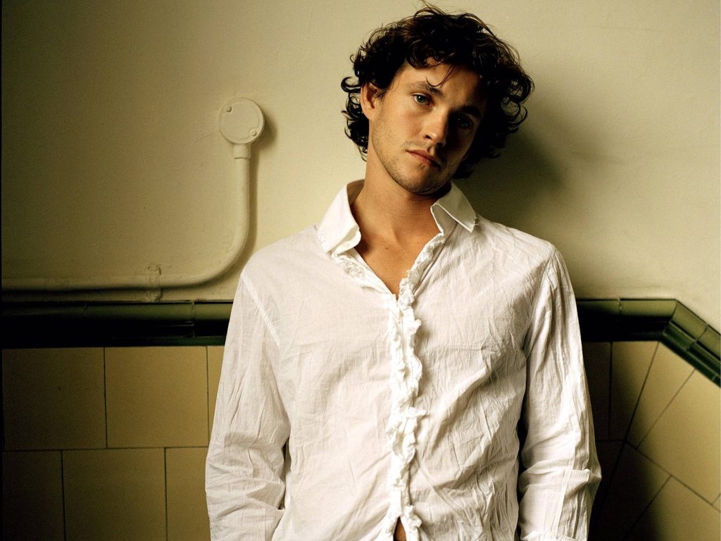 Hugh Dancy half-body portrait in white ruffled shirt against minimalist tiled wall background Wallpaper