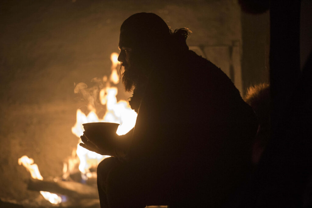 Hugh Glass from The Revenant seated by fire holding bowl intense battle scene wallpaper background