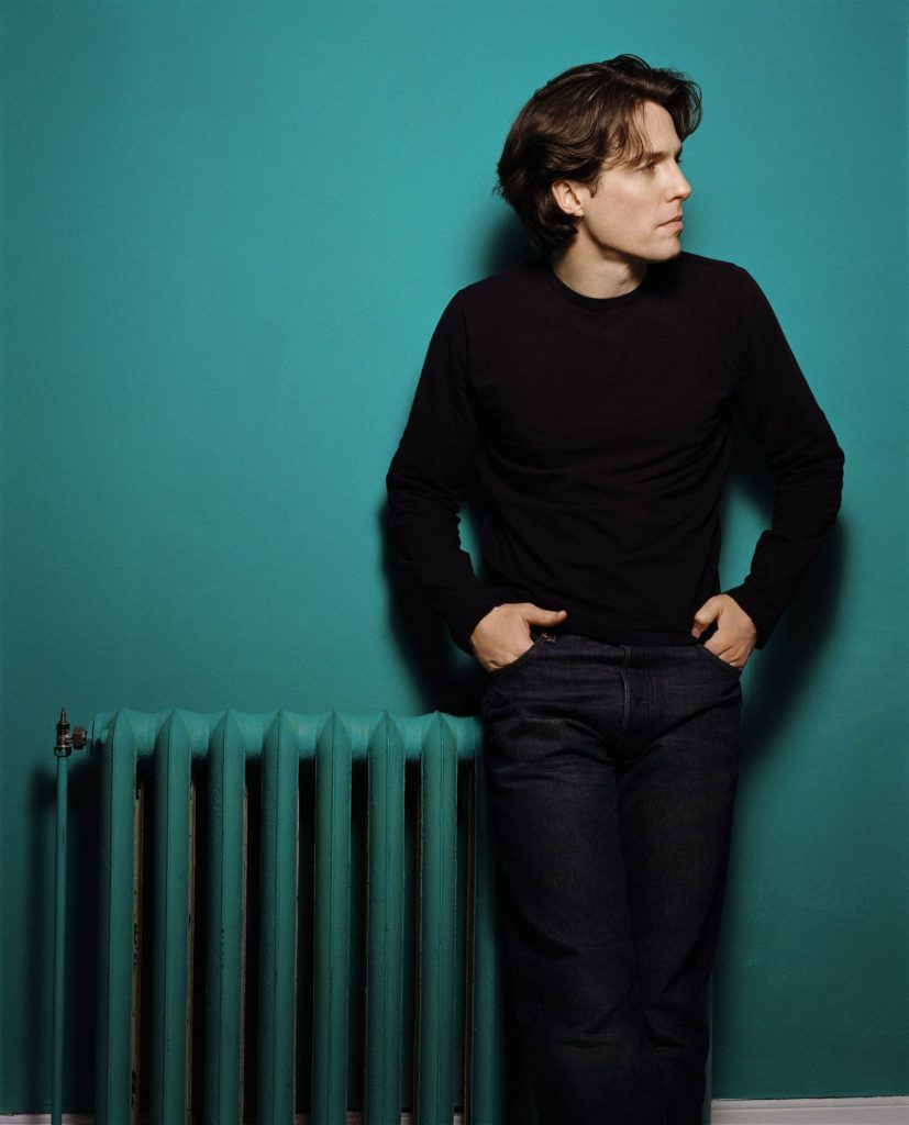 Hugh Grant portrait leaning on vintage radiator black shirt dark denim cool green background Wallpaper