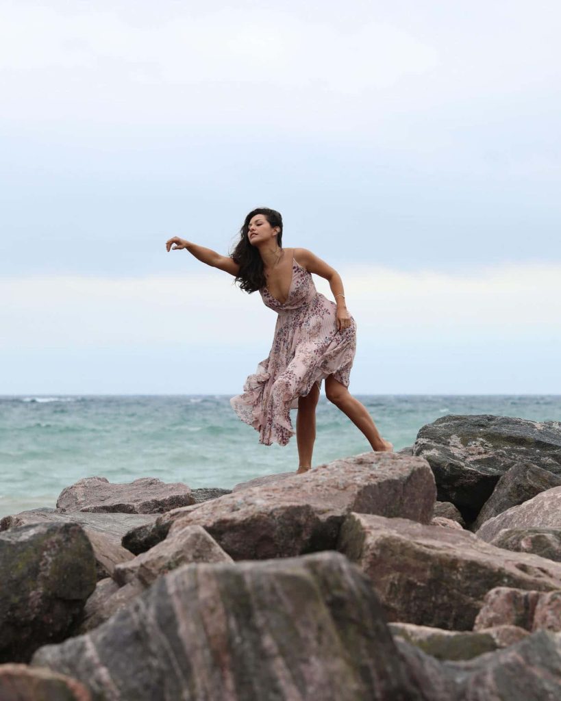 Actress Humberly González in flowing floral maxi dress posing on seaside rocks with ocean backdrop Wallpaper