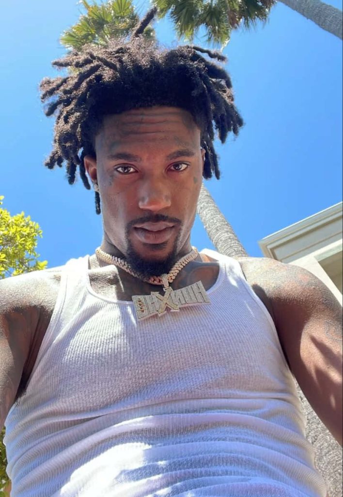 Rapper Hunxho confident selfie with dreadlocks diamond chain white tank top palm trees background Wallpaper