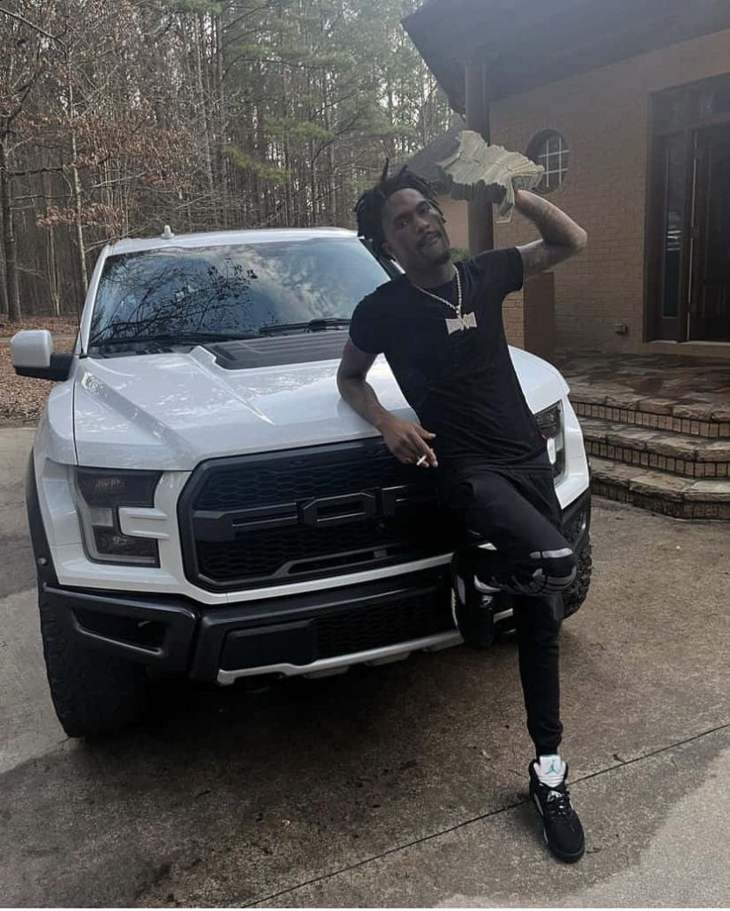 Rapper Hunxho in black outfit holding cash by white Ford F-150 Raptor in wooded driveway Wallpaper