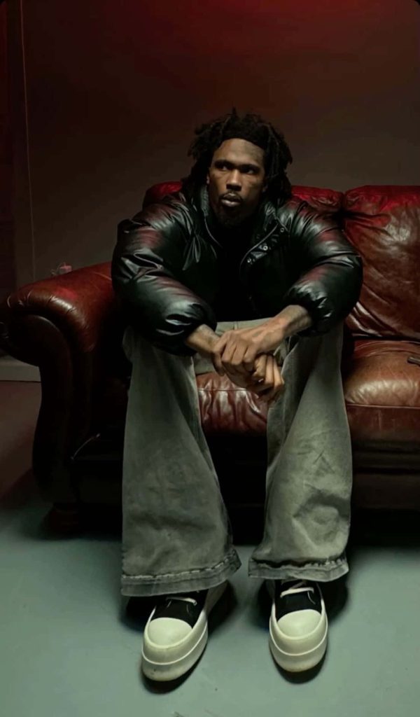 Moody portrait of rapper Hunxho on leather couch wearing black puffer jacket and denim Wallpaper