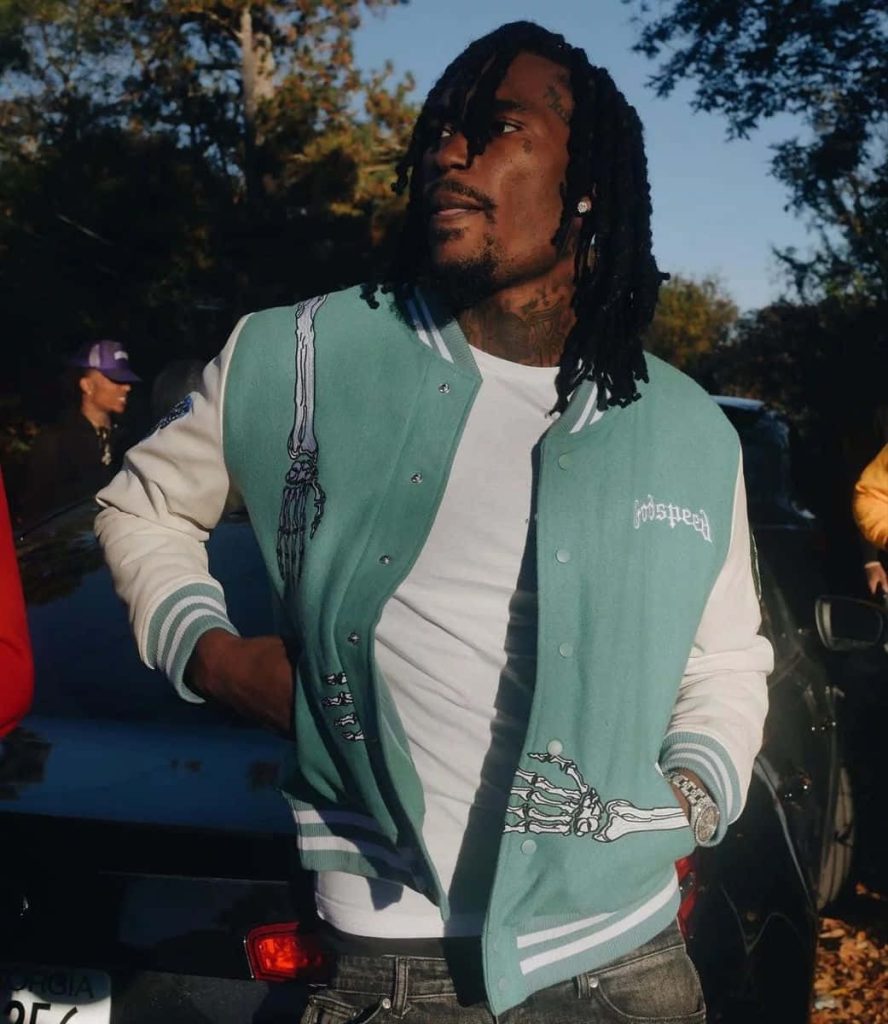 Rapper Hunxho outdoors in teal varsity jacket with skeleton hands urban style wallpaper background