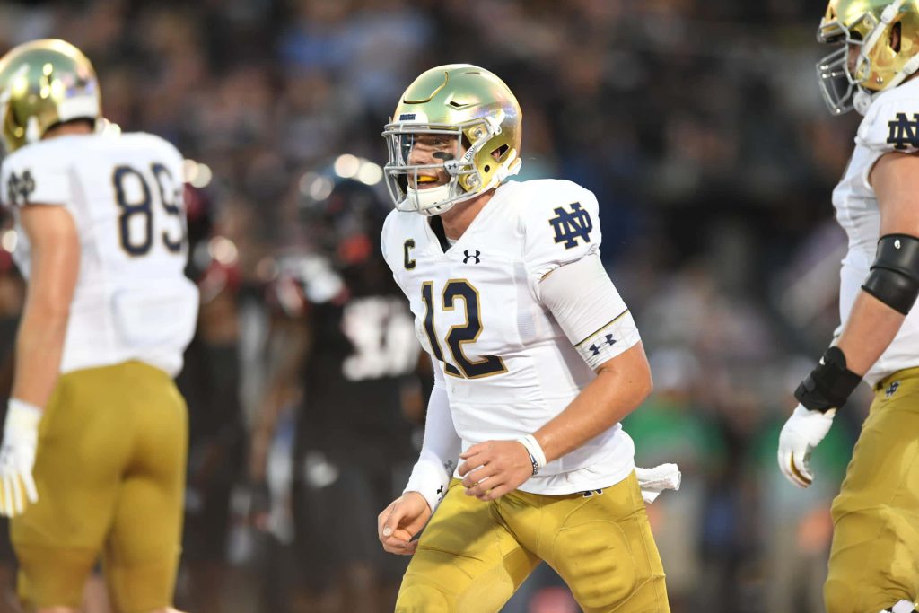 Notre Dame quarterback Ian Book in white and gold uniform intense collegiate gameplay wallpaper