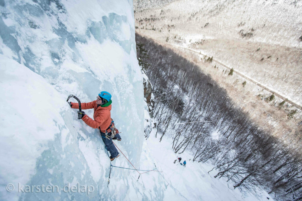 Climber in orange jacket with blue helmet ascends steep icy mountain using ice climbing gear Wallpaper