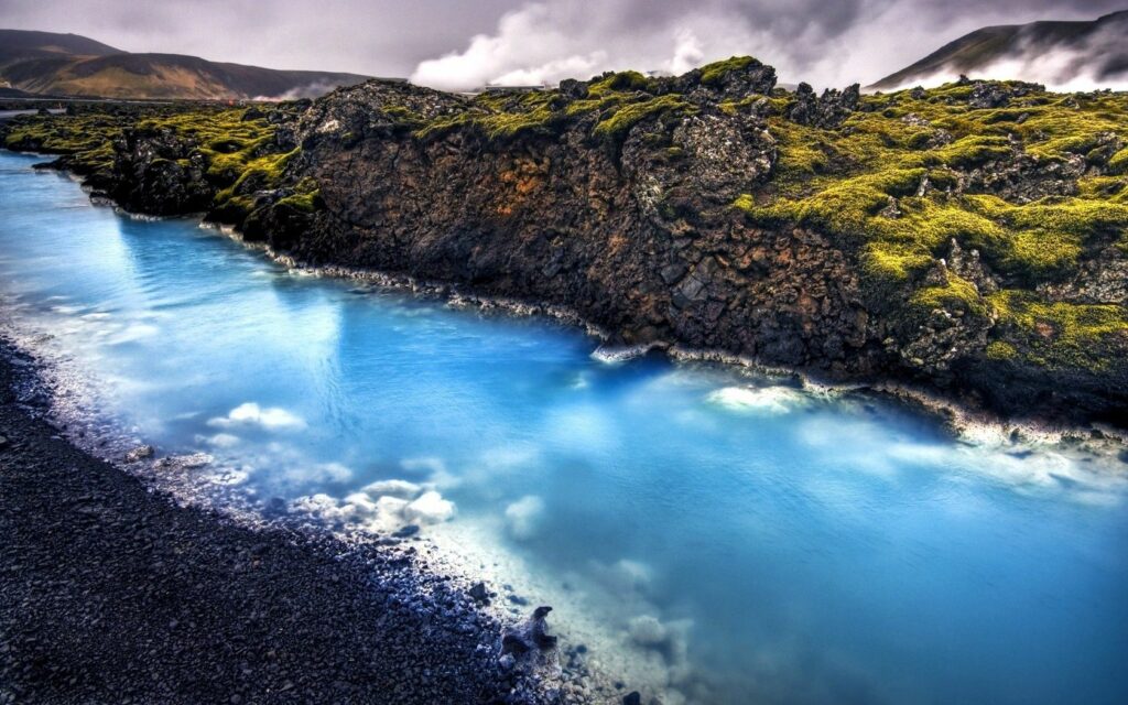 Icelandic hot spring river with bright blue water flowing past mossy stones and misty mountains Wallpaper