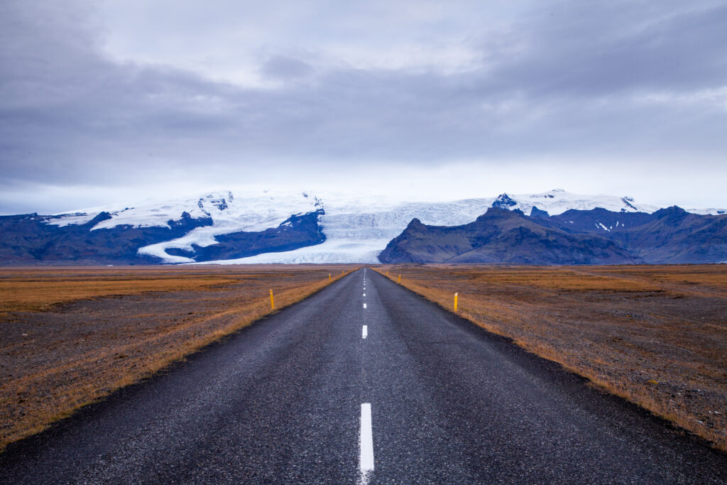 Straight asphalt road leads to snow-capped mountains under cloudy sky in rugged Iceland landscape Wallpaper
