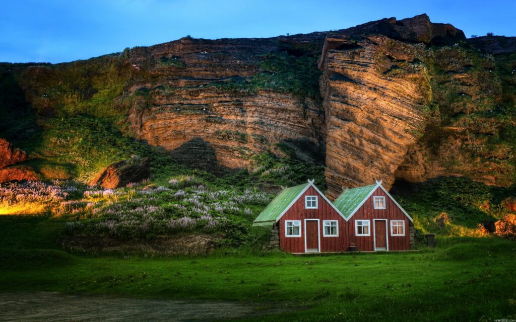 Red Icelandic lodges with green roofs by layered rock formations and soft meadow landscape Wallpaper