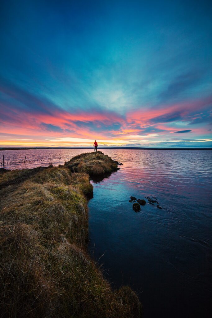 Icelandic sunset over tranquil lake with vibrant pink blue gold sky and lone figure on peninsula Wallpaper