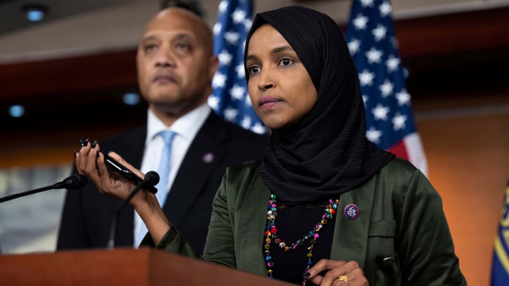 U.S. Representative Ilhan Omar in green blazer and hijab speaking at press conference with flags Wallpaper