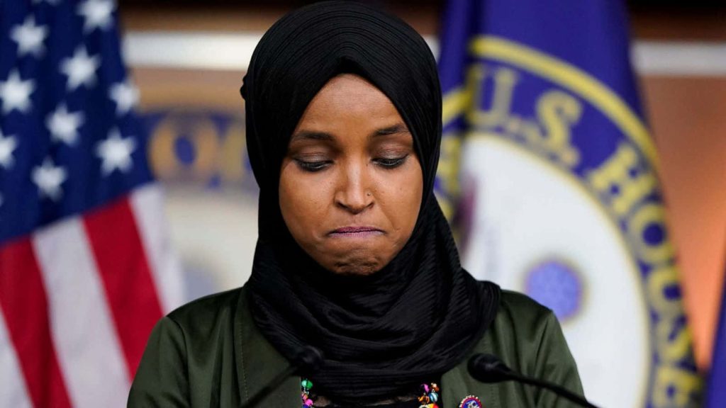 Pensive U.S. Representative Ilhan Omar in black hijab and olive blazer with House flags background Wallpaper