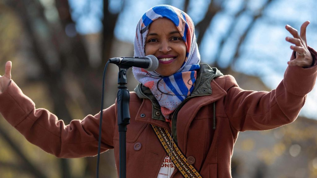 Ilhan Omar smiling with arms open wearing colorful hijab and brown coat at microphone outdoors Wallpaper