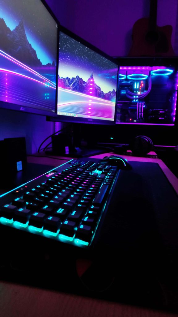 Cyberpunk gaming setup with dual monitors showing retro neon landscapes and RGB keyboard Wallpaper