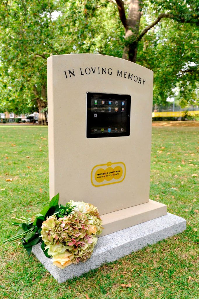 Modern In Loving Memory tombstone with iPad and flowers promoting charitable wills on green lawn Wallpaper