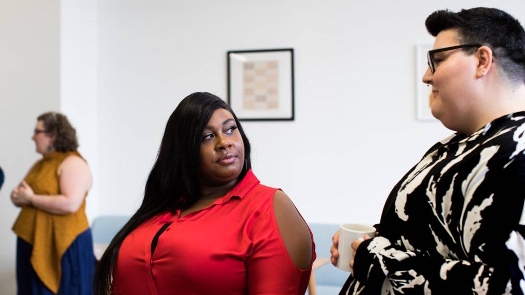 Black woman in red top sharing a knowing look with colleague holding mug in modern office Wallpaper