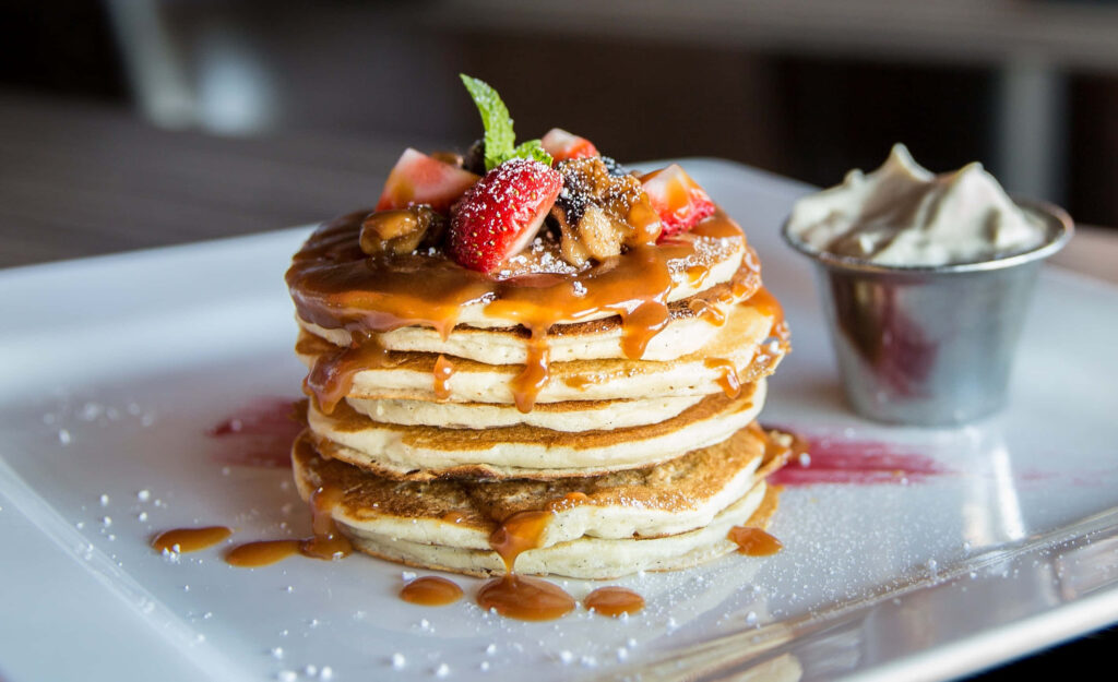 Delicious Pancake Paradise: A Tempting Stack of Fluffy Goodness, Drizzled in Rich Brown Syrup and Topped with a Dreamy Assortment of Irresistible Goodies Wallpaper