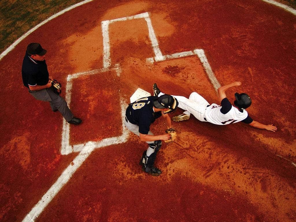 High-angle baseball play at home plate runner sliding past catcher with umpire watching intense game Wallpaper