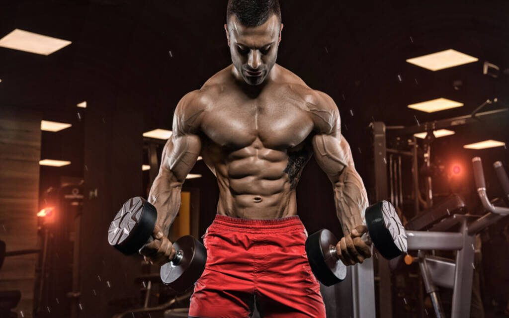 Muscular bodybuilder in red shorts doing dumbbell curls in dimly lit gym with dramatic lighting Wallpaper