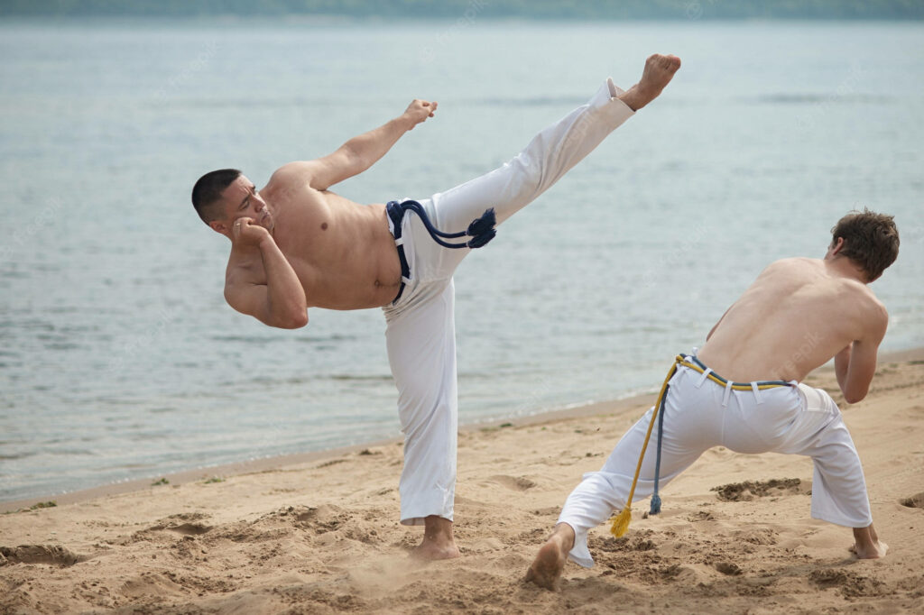 Two muscular men practicing intense capoeira with high kicks on sandy beach waterfront backdrop Wallpaper